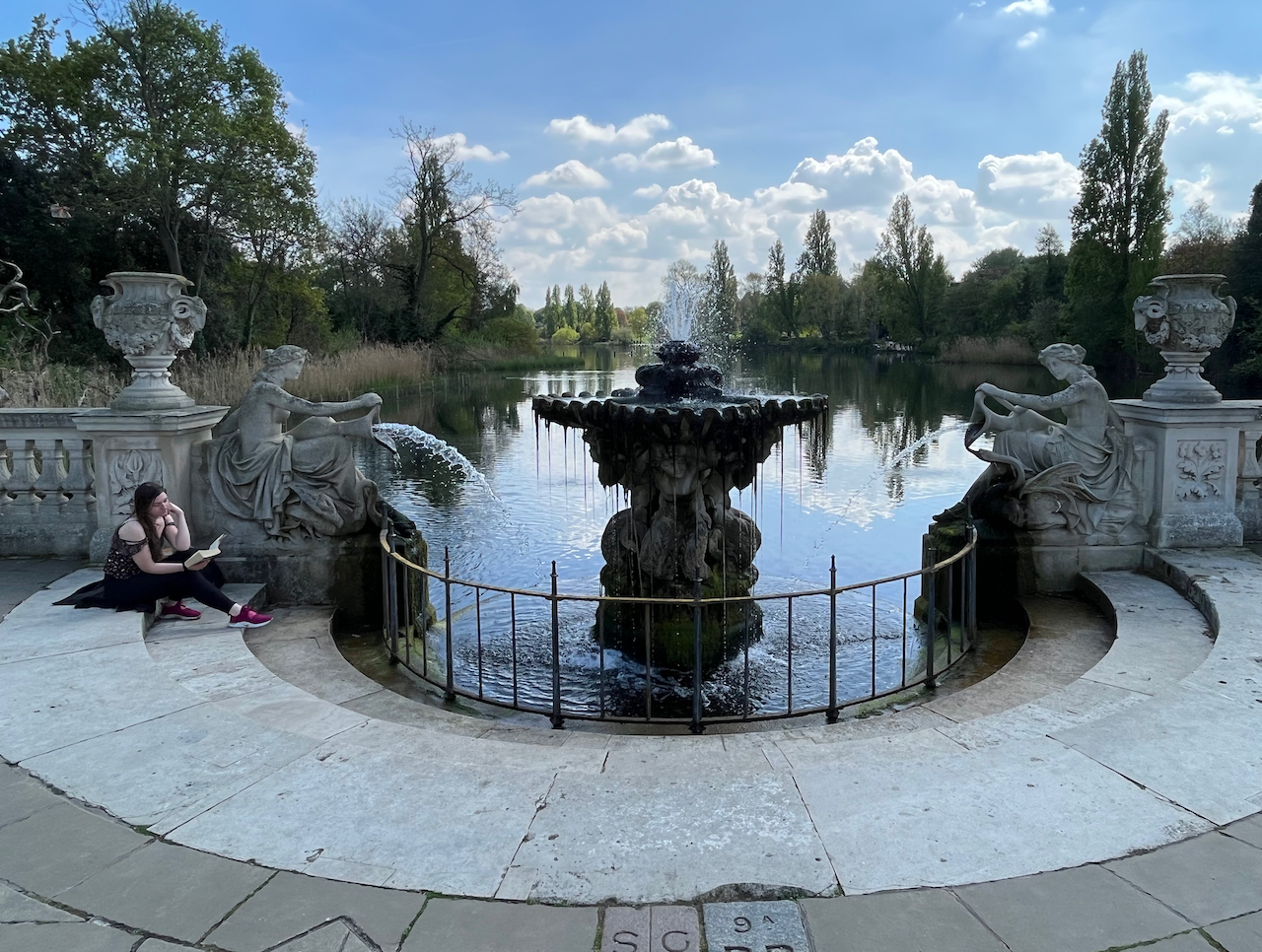 The Tazza fountain, an ornate pedestal with a fountain spraying out of the top, overlooking a lake called The Long Water, that flows around a corner in the distance towards the Serpentine in Hyde Park. On each side of the fountain is a statue of a seated woman pouring water out of a large urn towards the base of the central pedestal.