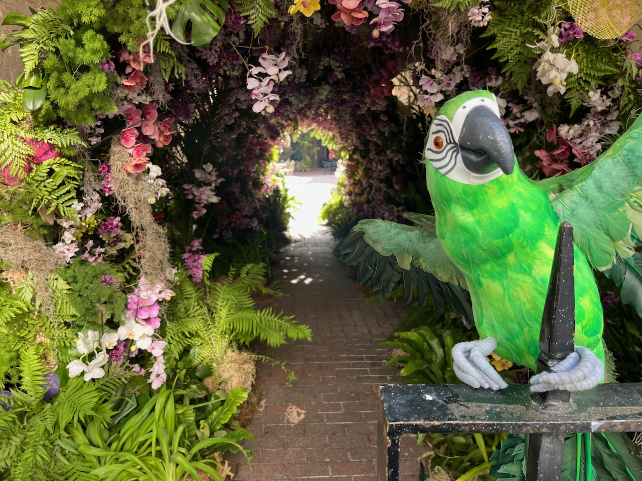 A model of a parrot, with a green body, white face and a large black beak, clinging on to some black railings with its light grey claws, with a tunnel made of greenery and colourful orchids in the background.