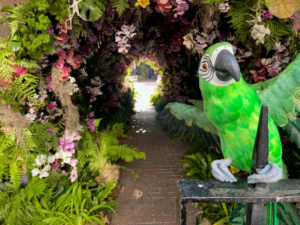 A model of a parrot, with a green body, white face and a large black beak, clinging on to some black railings with its light grey claws, with a tunnel made of greenery and colourful orchids in the background.