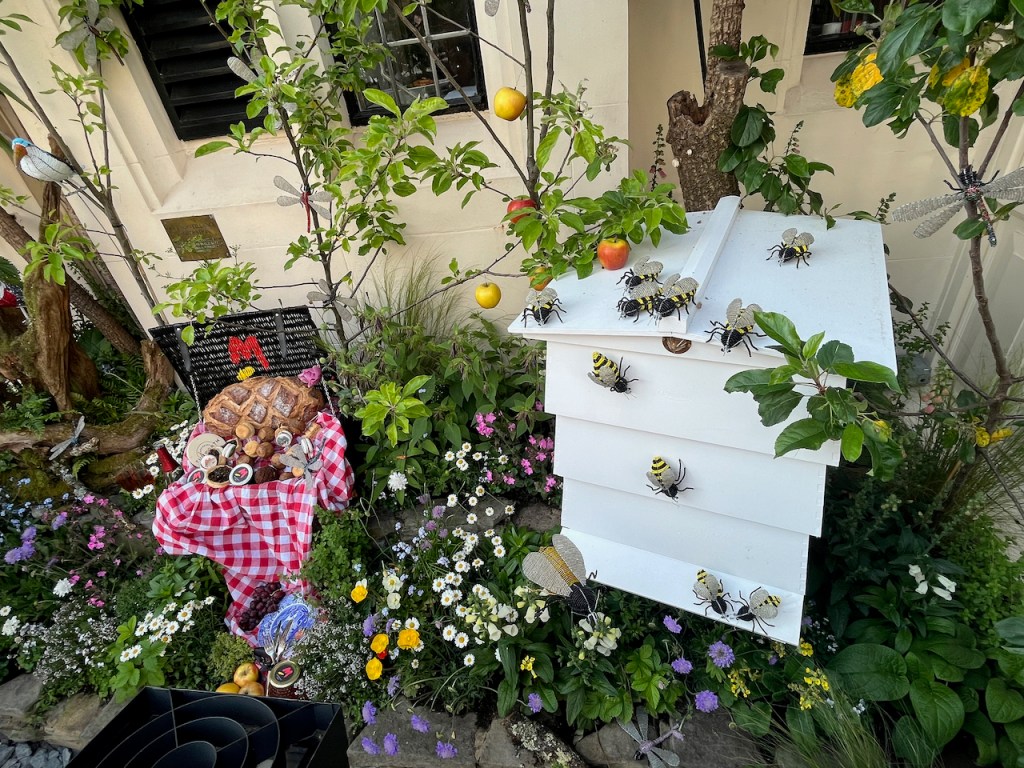 Floral installation with lots of colourful flowers, some bees climbing over a white beehive, and a picnic hamper with meaty items and other treats.