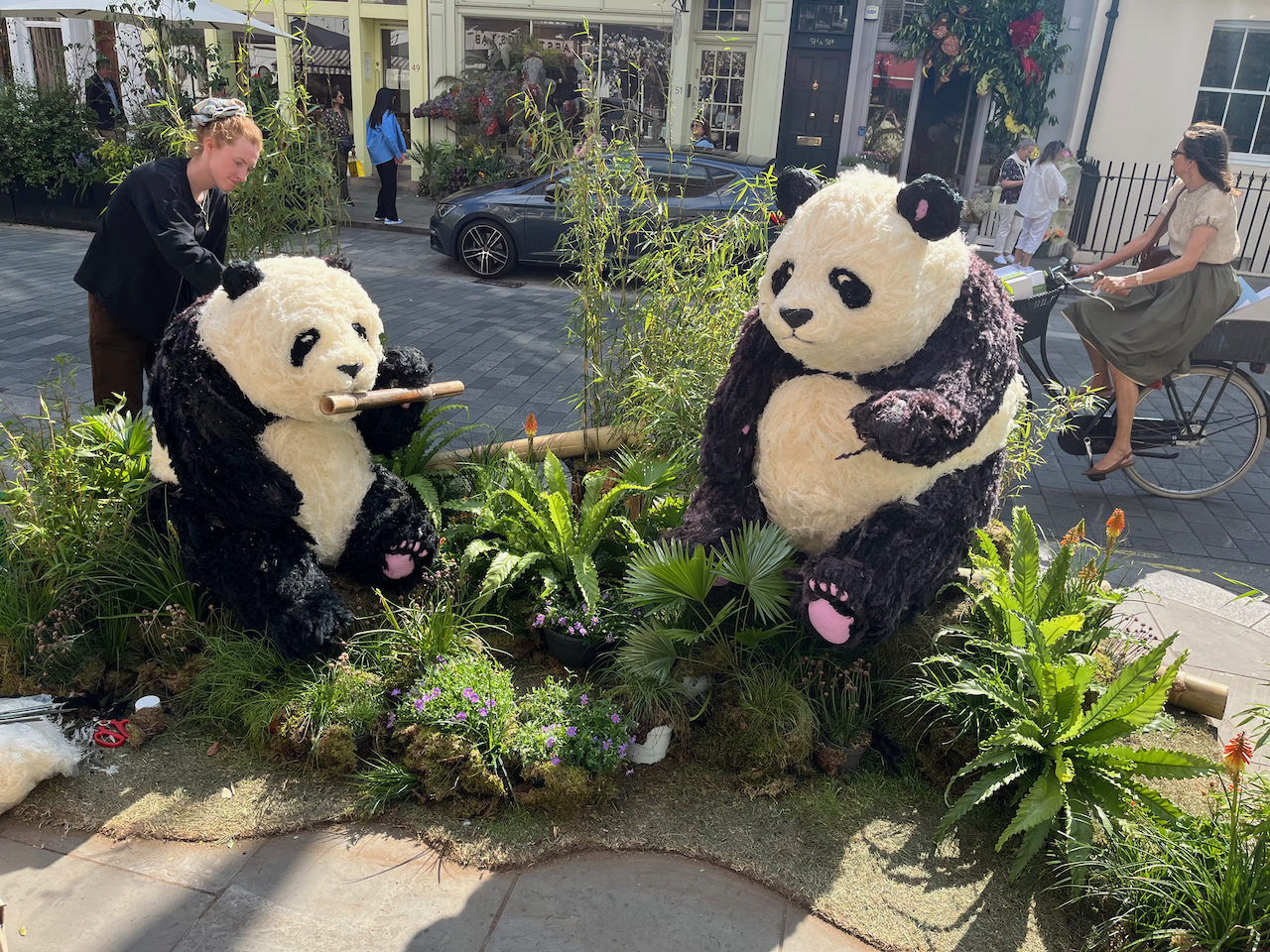 Floral installation featuring 2 large black and white pandas, one eating a thick piece of bamboo cane, as they sit amongst greenery and small blue flowers.