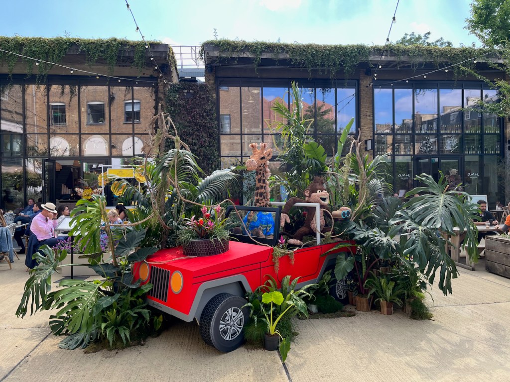 In amongst a lot of green leafy plants, a giraffe drives a red jeep, while a monkey with a big smile on its face holds a pair of binoculars as it sits in the back looking around.