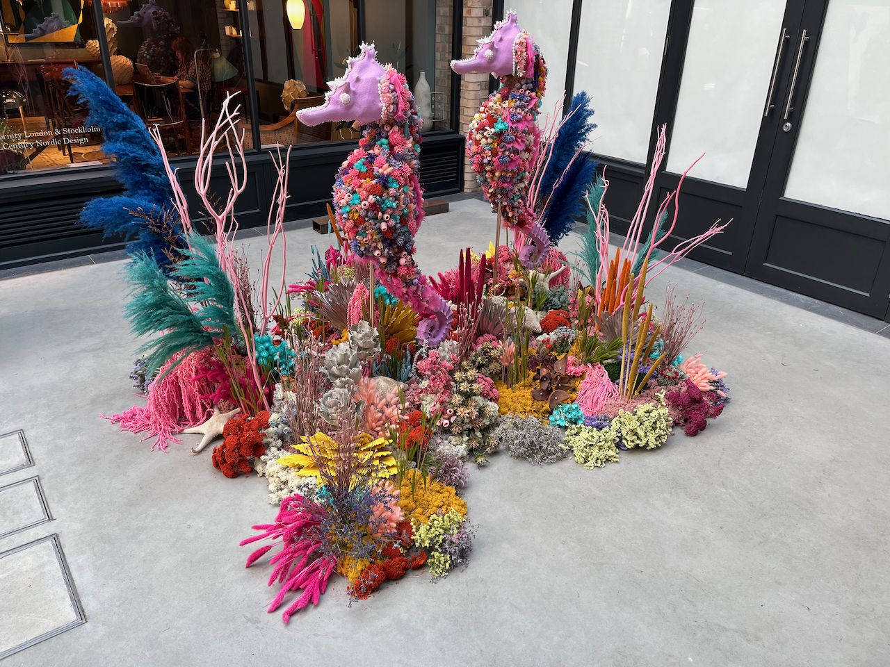 Models of 2 pink seahorses decorated in colourful flowers, one behind the other and facing the same direction, standing in a bed of colourful flowers.