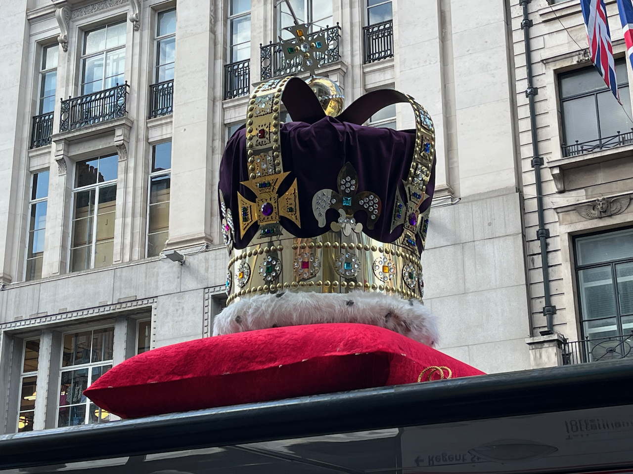 An oversized crown, with a soft purple fabric in the centre surrounded by gold and jewel encrusted detailing, sitting on a red cushion, on the roof of a bus stop in Oxford Street.