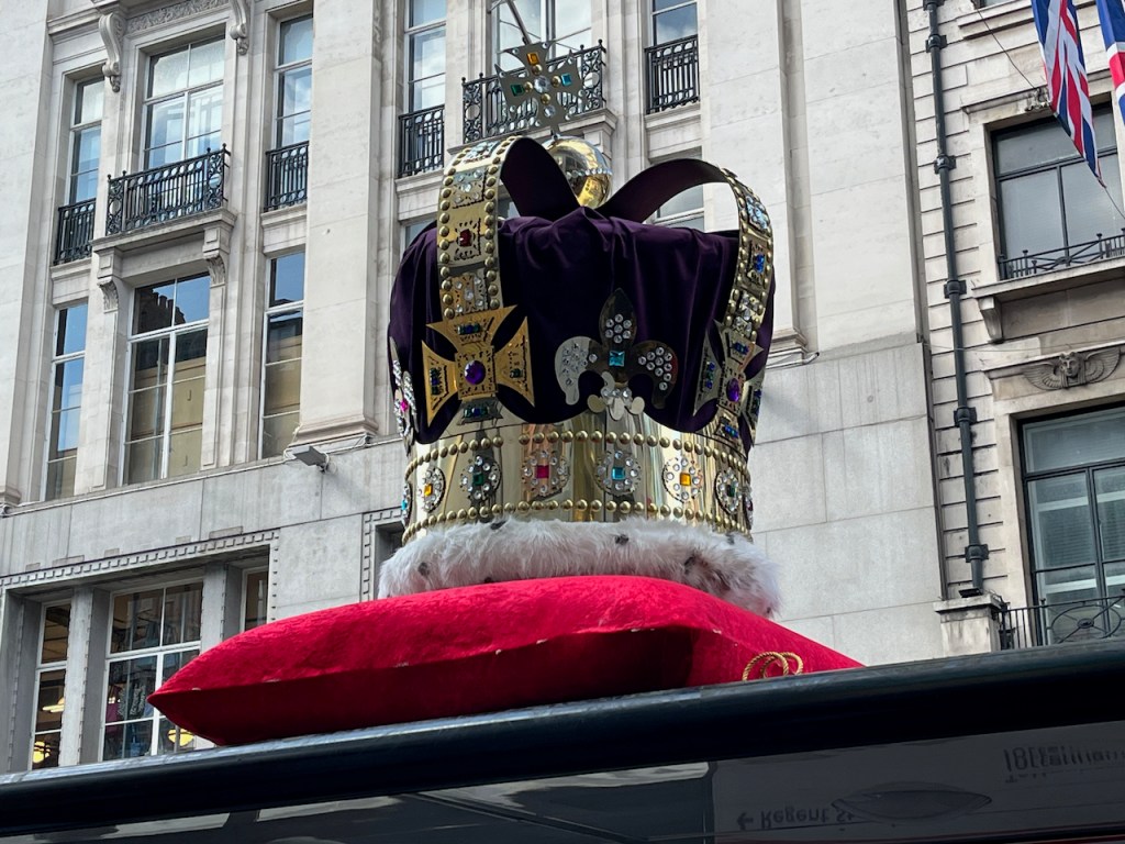 An oversized crown, with a soft purple fabric in the centre surrounded by gold and jewel encrusted detailing, sitting on a red cushion, on the roof of a bus stop in Oxford Street.