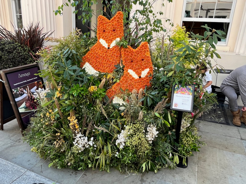 Floral installation featuring 2 foxes. The foxes are represented in a 2-dimensional way, facing us with their heads and chests visible among the white flowers and greenery.
