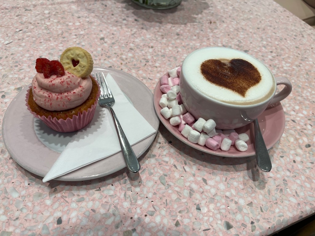 A cupcake with pink icing swirled neatly on the top, and a mini Jammie Dodger embedded into it. The Dodger is a round biscuit with a red jam-infused heart in the centre. Next to it is a mug of hot chocolate, with a big chocolate heart in the centre of the white foamy surface, plus lots of pink and white mini marshmallows in the saucer around it.