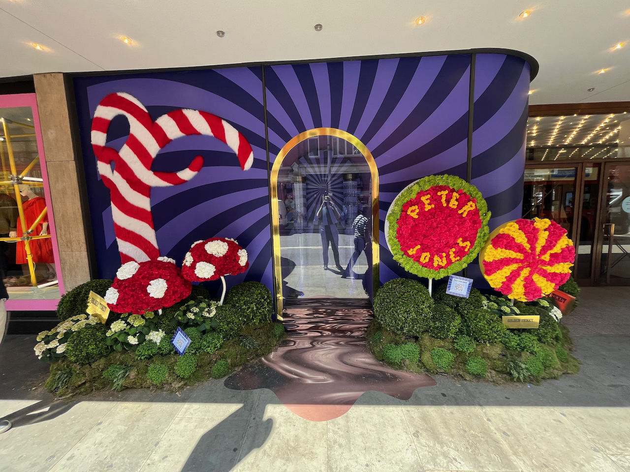 Floral display representing Charlie And The Chocolate Factory, including an oversized red and white striped candy cane, 2 large toadstools that are red with white spots, and 2 large round lollipops, one of which has a red and yellow spiral on it, and the other has the words Peter Jones in yellow lettering against a red background and a green border. There are also a few golden ticket envelopes nestled into the greenery around the large sweets.