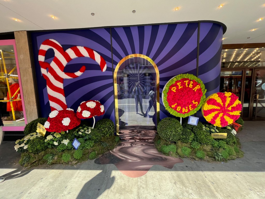 Floral display representing Charlie And The Chocolate Factory, including an oversized red and white striped candy cane, 2 large toadstools that are red with white spots, and 2 large round lollipops, one of which has a red and yellow spiral on it, and the other has the words Peter Jones in yellow lettering against a red background and a green border. There are also a few golden ticket envelopes nestled into the greenery around the large sweets.