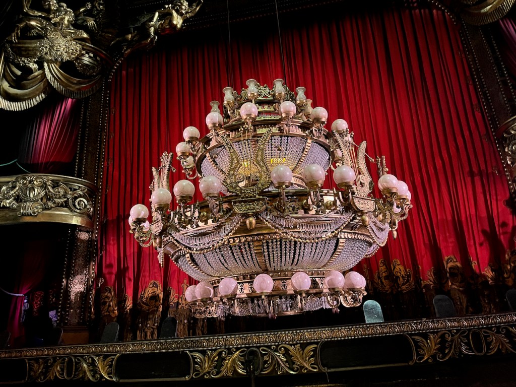 The huge, ornate chandelier, seen in the interval, with white electrical bulbs around the top, centre and bottom of its globe-like structure, the rest of the space filled by chains of crystals and gold decorations.