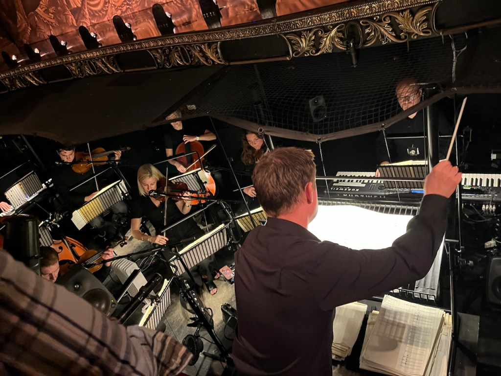 A man conducting a small orchestra at the Phantom Of The Opera. Here we see the left side of the orchestra pit, including strings players and a keyboard player.
