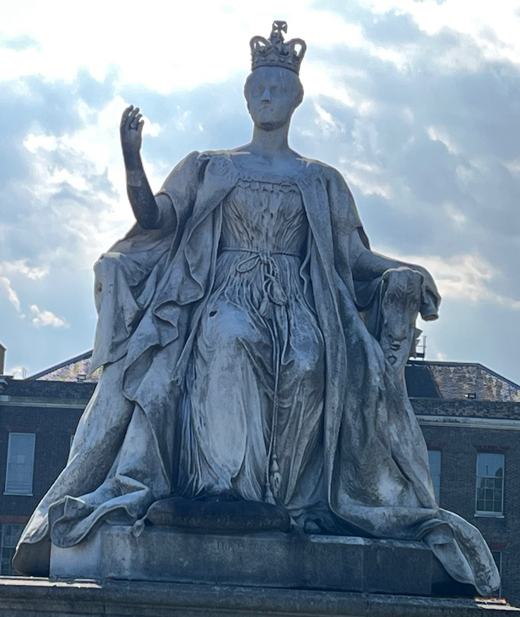 A white stone statue of Queen Victoria, who is seated with her right arm bent at the elbow so her hand is raised, and she is wearing a crown on her head.
