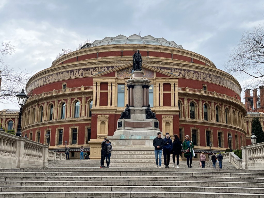The huge ellipse shaped Royal Albert Hall building, with lots of ornate gold decorations surrounding the red brick work. In front of the hall, at the top of some very wide steps, is The Memorial to the Great Exhibition, consisting of a tall stone plinth, with figures sat on each side of the stone column in its centre, and on the top of the column is a statue of Albert, Prince Consort.