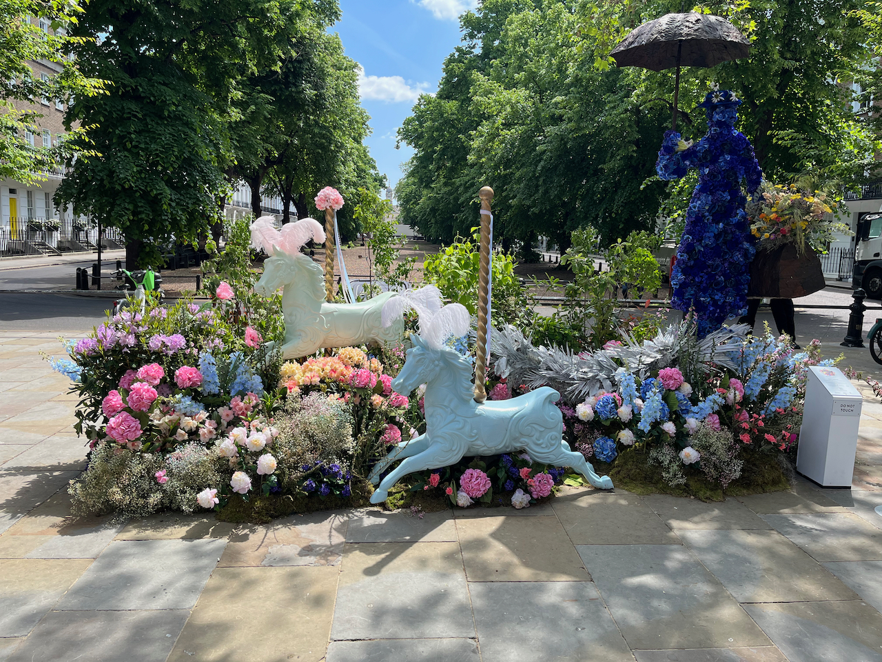A floral display representing Mary Poppins, who is represented as a tall dark blue figure holding a black umbrella. Two horses from a fairground carousel ride, complete with the twisting metal poles sticking up out of their backs, are paused in mid air in front of her, as if an invisible carousel ride is still running. The rest of the display around them is filled with flowers in a variety of bright colours, including whites, blues and pinks.