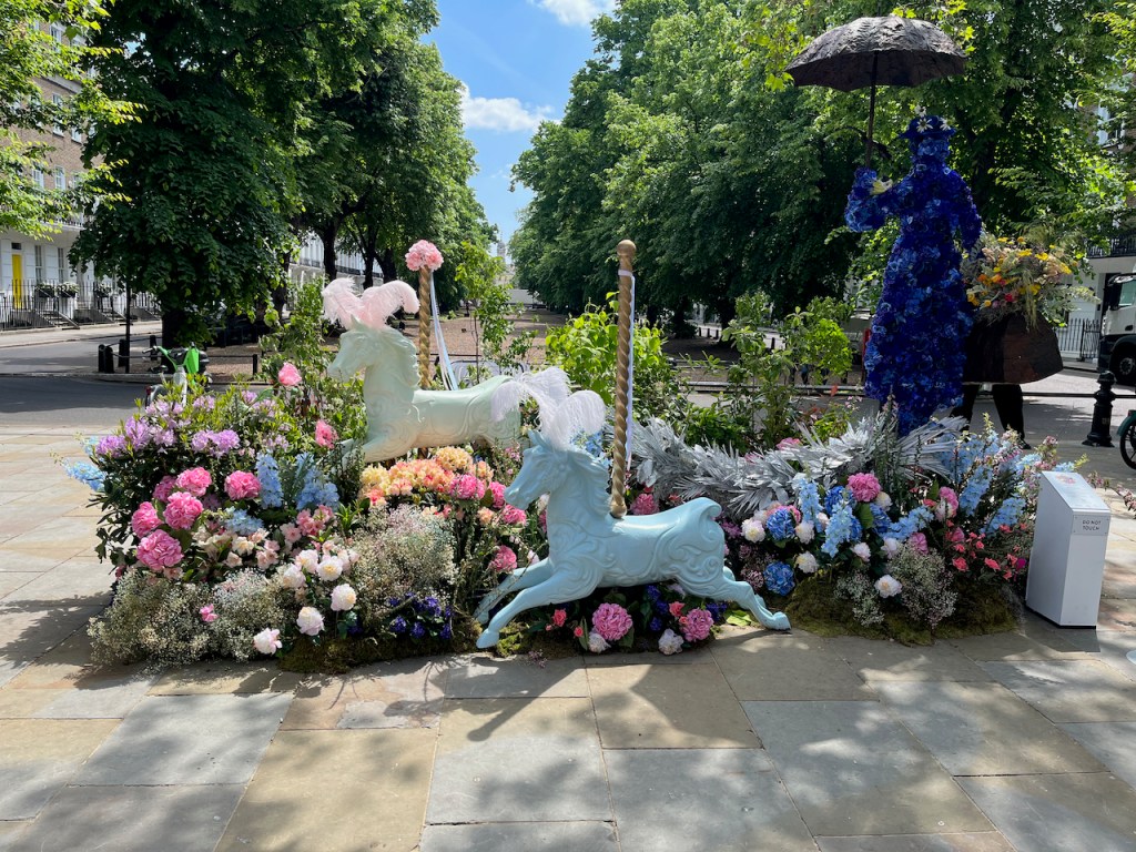 A floral display representing Mary Poppins, who is represented as a tall dark blue figure holding a black umbrella. Two horses from a fairground carousel ride, complete with the twisting metal poles sticking up out of their backs, are paused in mid air in front of her, as if an invisible carousel ride is still running. The rest of the display around them is filled with flowers in a variety of bright colours, including whites, blues and pinks.