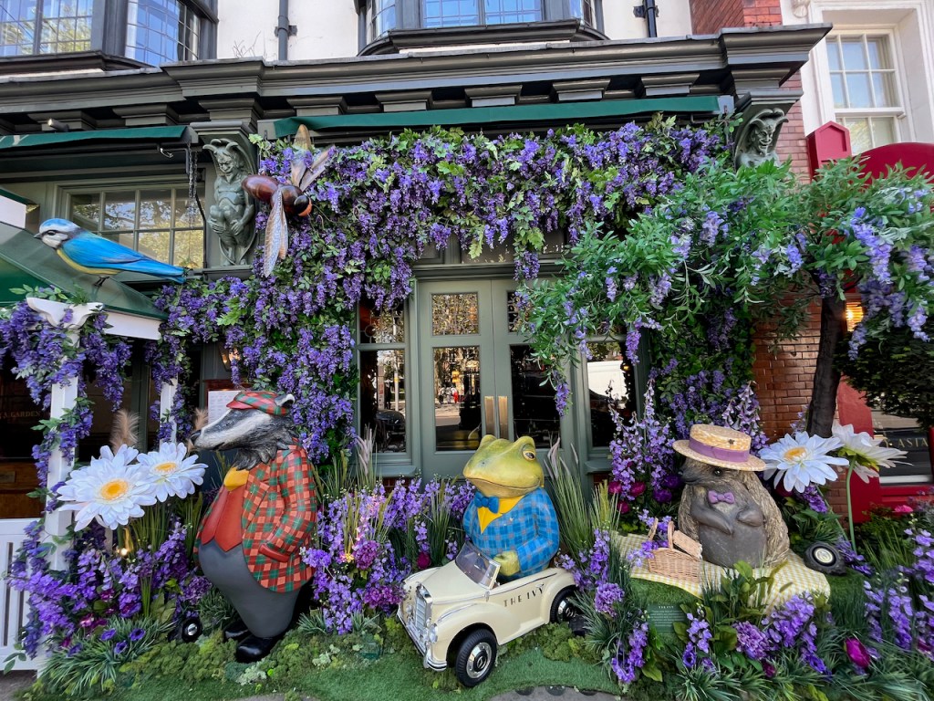 A large display outside The Ivy in King's Road, Chelsea. Lots of purple flowers and greenery are spread around the windows and on the ground, surrounding models of Badger, Toad and Rat from Wind In The Willows. Models of flying insects and a blue tit can also be seen amongst the flowers above them.
