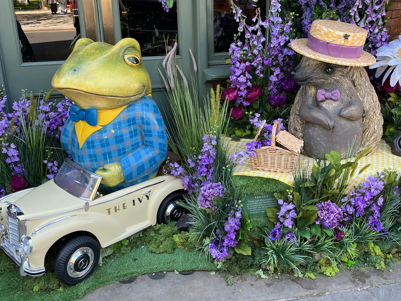 Close-up of Toad and Rat, surrounded by purple flowers and greenery. Toad is dressed in a blue checked jacket over a yellow shirt with a blue bow tie, and the lower half of his plump figure only just squeezes into the small white car he's sat in. Next to him, Rat wears a straw boater hat and a small purple bow tie, while sitting on a picnic blanket with a wicker picnic basket.
