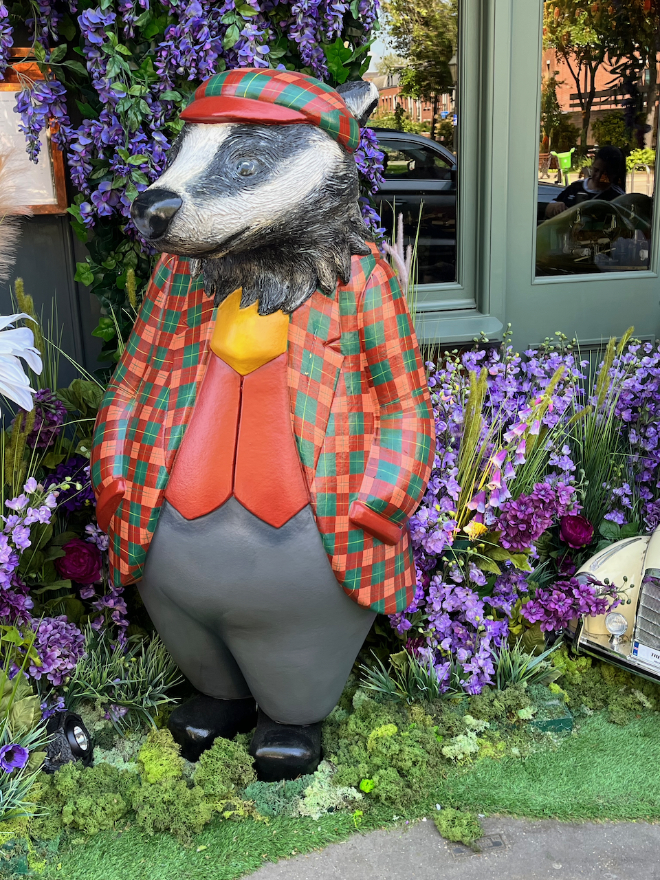 Close-up of Badger in The Ivy's Wind In The Willows display, who is wearing a jacket and a flat cap that both have a red and green check design. Beneath his jacket he is also wearing a yellow shirt, and on his plump legs he's wearing grey trousers and black shoes.