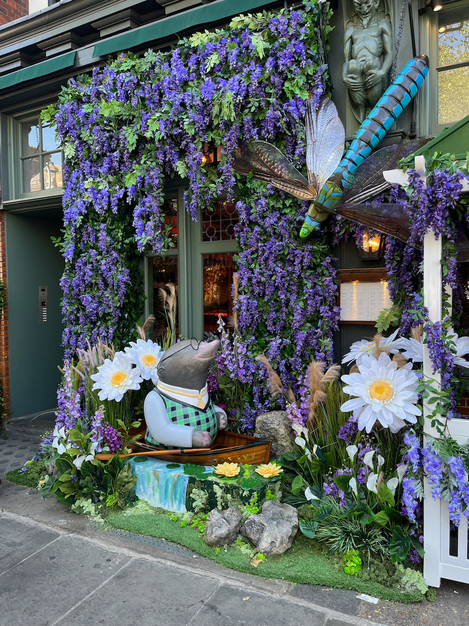 The other half of The Ivy's Wind in The Willows display, showing Mole in a wooden boat. He too is surrounded by purple flowers hanging around the windows, but there are also a few bigger flowers with white petals and yellow centres in front and behind him. A large model of a colourful flying insect, like a dragonfly, is among the foliage above him.