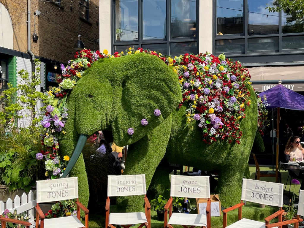 A large green model of an elephant, which looks as if it's made out of turf. Its back is covered in lots of colourful flowers, and there are also 3 pink flowers attached to one ear, like piercings. The elephant is nicknamed Jumbo Jones, while chairs in front of it also have names on the back, including Quincy Jones, Indiana Jones, Grace Jones and Bridget Jones.