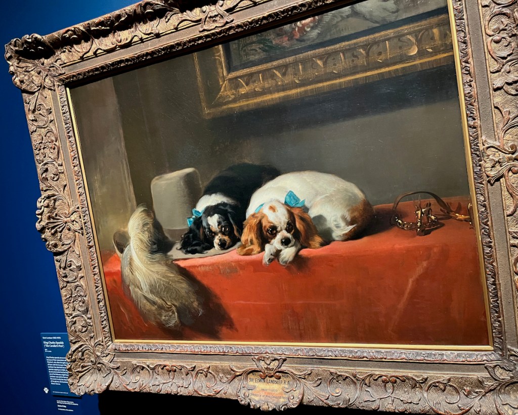 A painting of two King Charles Cavalier Spaniels, one with black and white fur, the other with brown and white fur, laying down on their fronts, on a table covered with a red cloth.