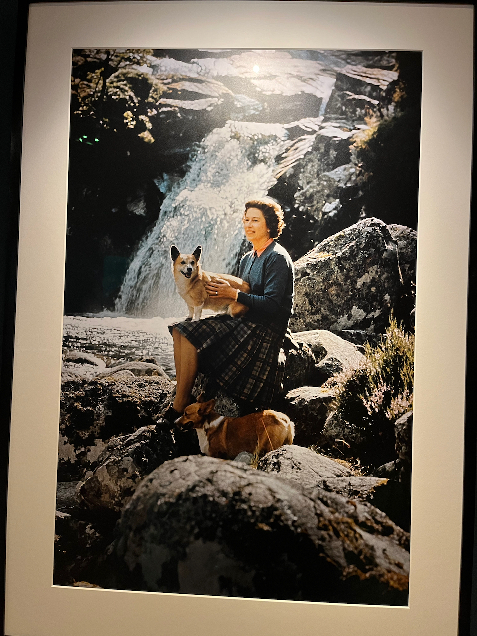 A photo of Queen Elizabeth the Second as a young adult, sitting on a rock in front of a waterfall, with a smiling Corgi dog standing on her lap.