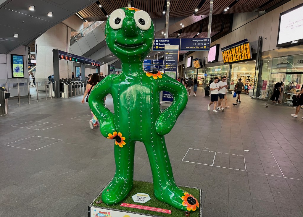 Cactus Morph in London Bridge station. This is a green statue of Morph with white spikes drawn all over him, along with occasional flowers with yellow and orange petals and black centres.