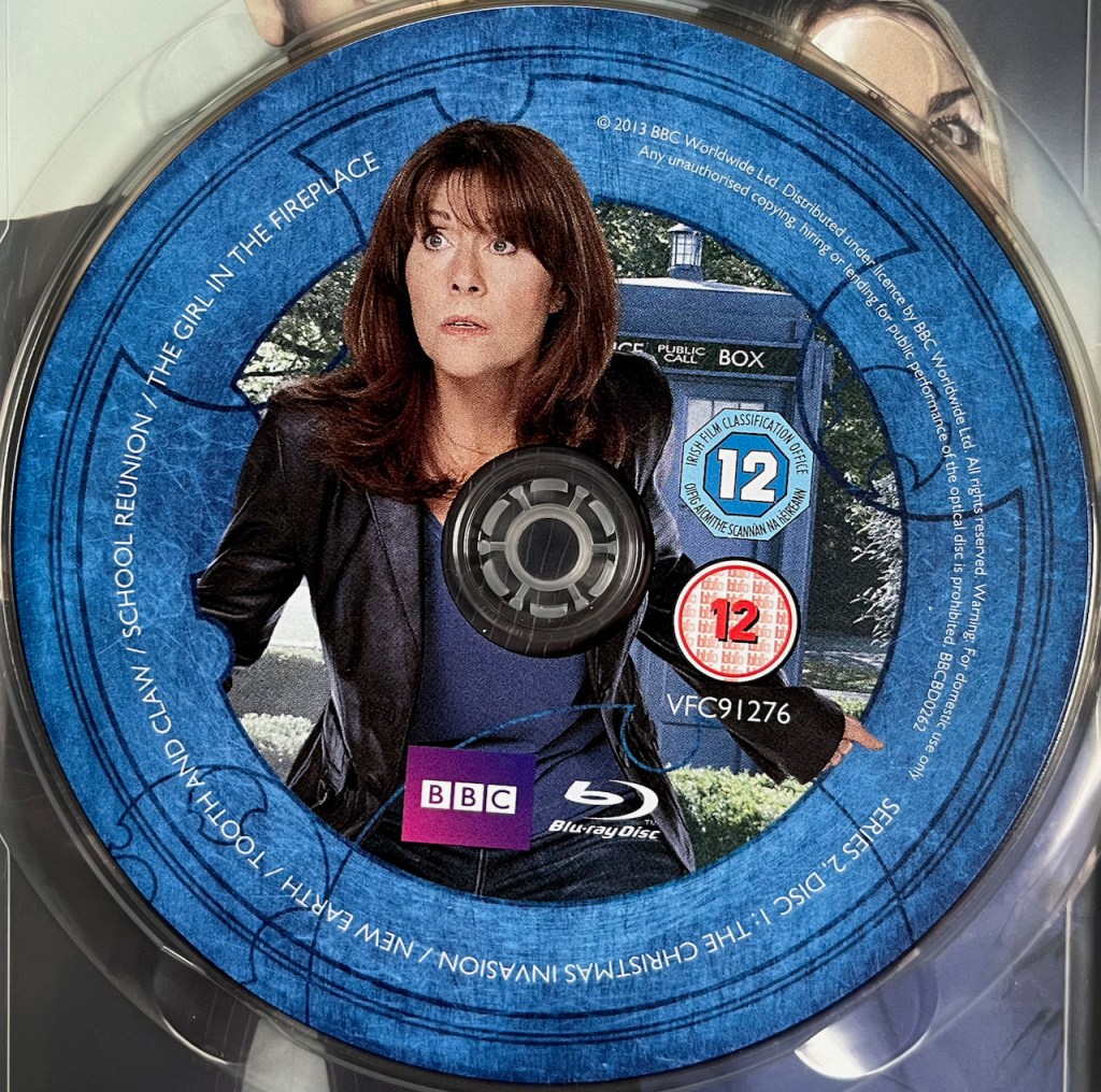 Disc 1 in the Doctor Who Series 2 Blu-ray set, featuring a photo of Sarah Jane Smith wearing a leather jacket over a blue top. A blue border around the edge of the disc contains the episode names and copyright information.