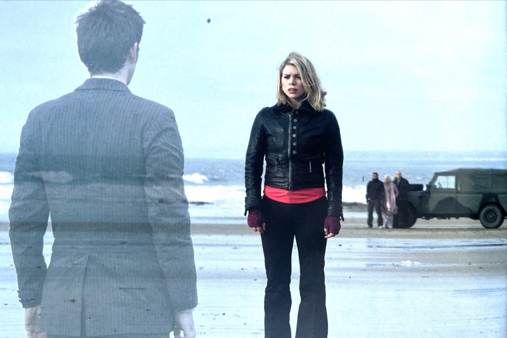 Image from the Doctor Who episode Doomsday, featured in the DVD booklet for Series 2, showing Rose standing on a beach as she looks at a semi-transparent hologram of David Tennant's Doctor. In the background, Mickey, Jackie and Pete watch as they stand next to a green jeep.