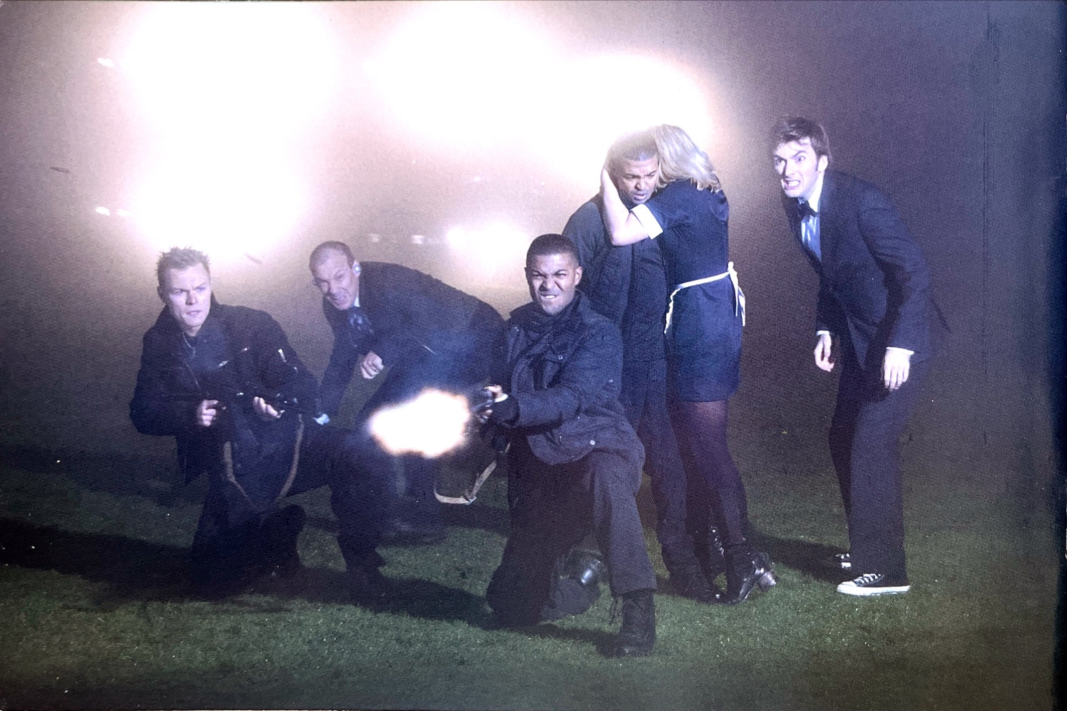 Image from the Doctor Who episode Rise Of The Cybermen, featured in the DVD booklet for Series 2. It shows David Tennant's Doctor and his companions as they confront the approaching Cybermen. Of the primary characters, the Doctor in his suit and bow tie looks angry, while Rose is dressed in a maid's apron and has her back to the action while she hugs Mickey. Next to them are 3 characters from the parallel world. Mickey's doppelganger Ricky is firing a gun, while his friend Jake also has a gun pointed ahead of him, and Pete Tyler looks on in fear.