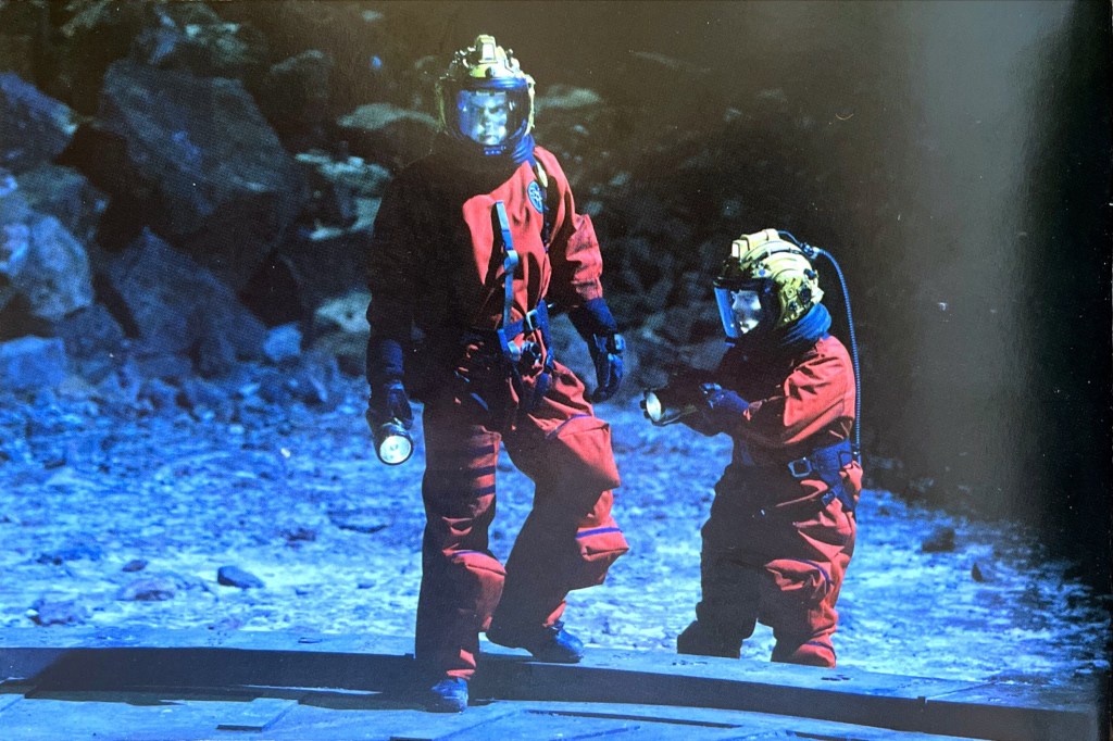 Image from the Doctor Who episode The Satan Pit, featured in the DVD booklet for Series 2, showing David Tennant's Doctor and another character wearing red space suits, black gloves and yellow space helmets with clear visors, as they hold torches and carefully explore a rocky landscape.