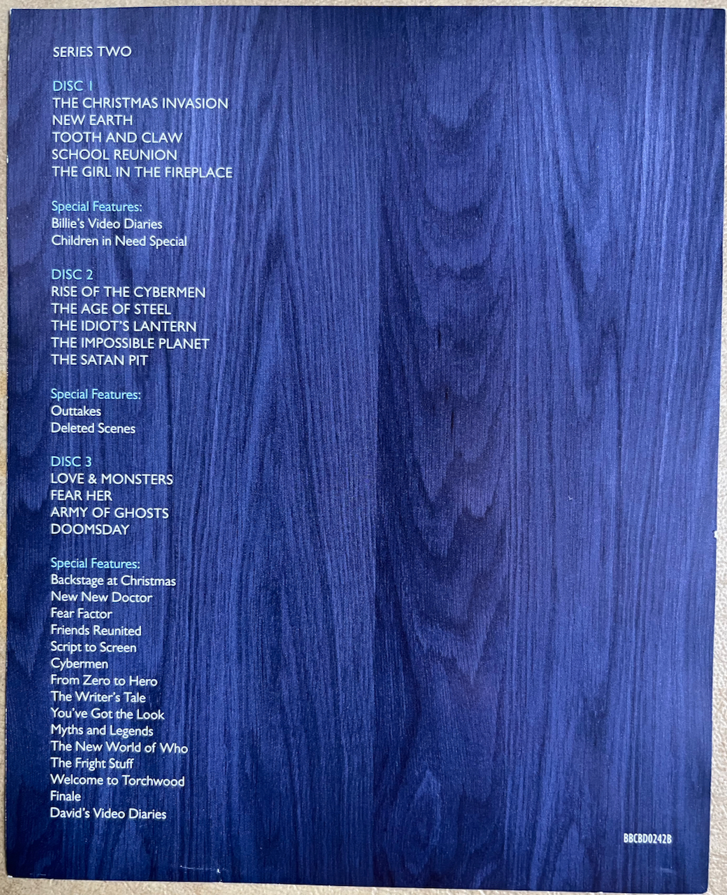 Rear side of the flyer included with the Doctor Who Series 2 Blu-ray Steelbook, showing the list of episodes and extras on each of the 3 discs, in white text over a close-up of the blue wood that makes up the exterior of the Tardis.
