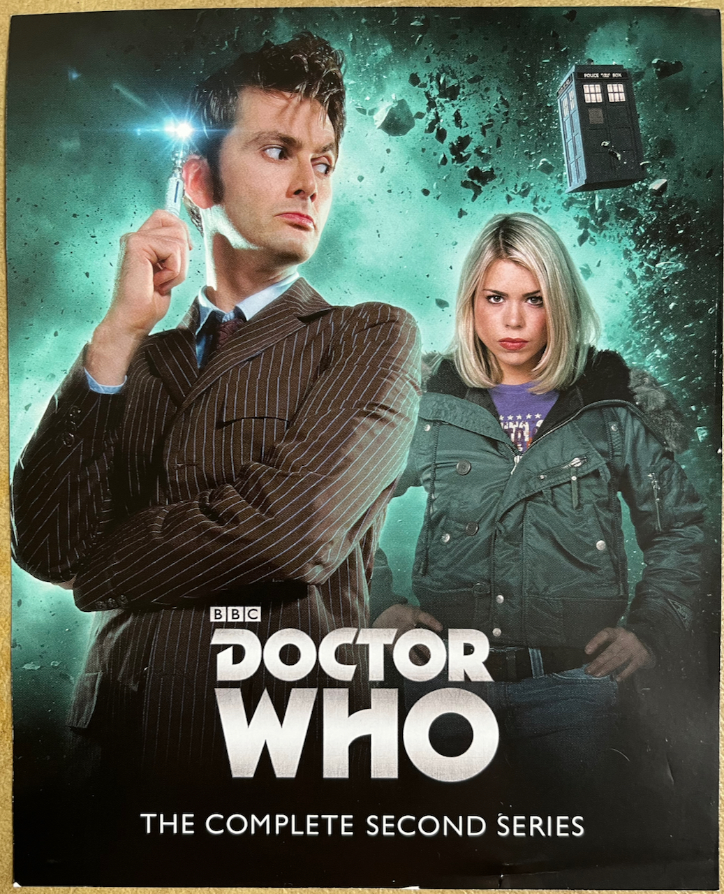 Front side of the flyer included with the Doctor Who Series 2 Blu-ray Steelbook, featuring a photo of David Tennant's Doctor wearing a pinstripe suit and holding up his sonic screwdriver, while looking back at Rose who is wearing a green jacket with her hands on her hips. In the background, the Tardis flies through an explosion of rocks above them.
