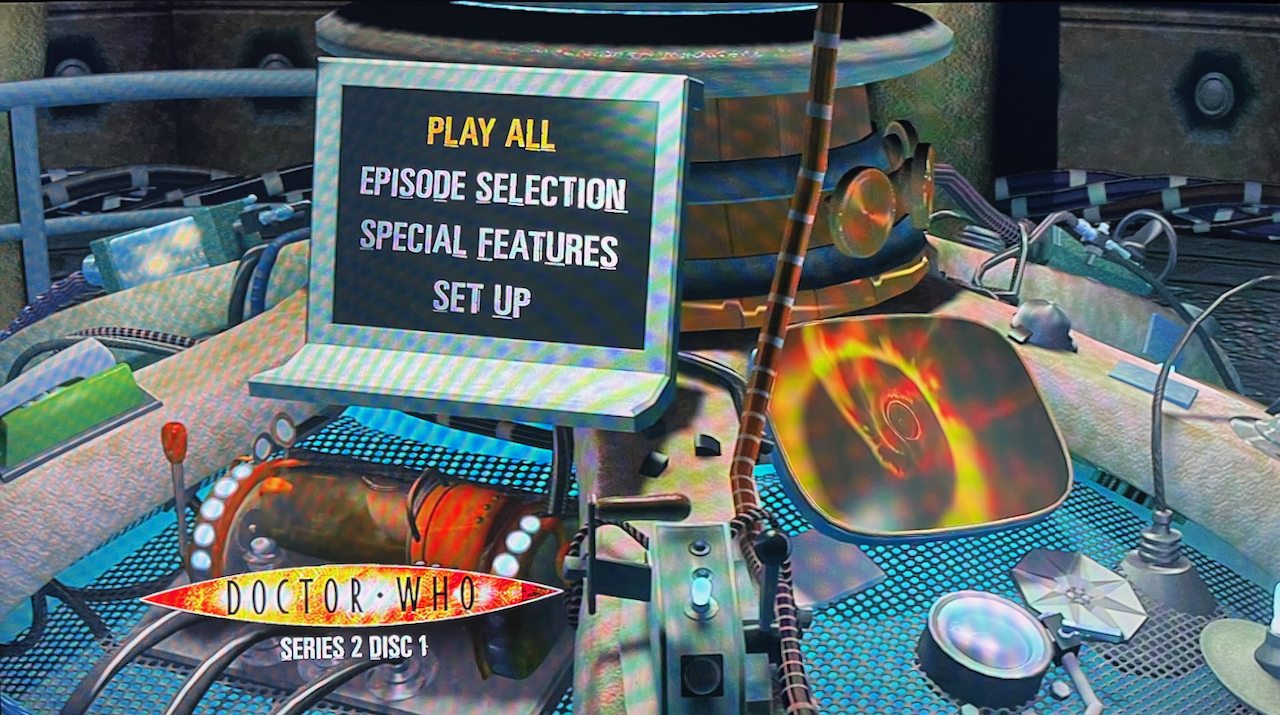 Blu-ray menu for Doctor Who Series 2. The 4 options - Play All, Episode Selection, Special Features and Set Up - are displayed on a computer screen on the Tardis console, with various lights, levers and buttons around it, and another screen to the right with a view of the swirling time vortex.