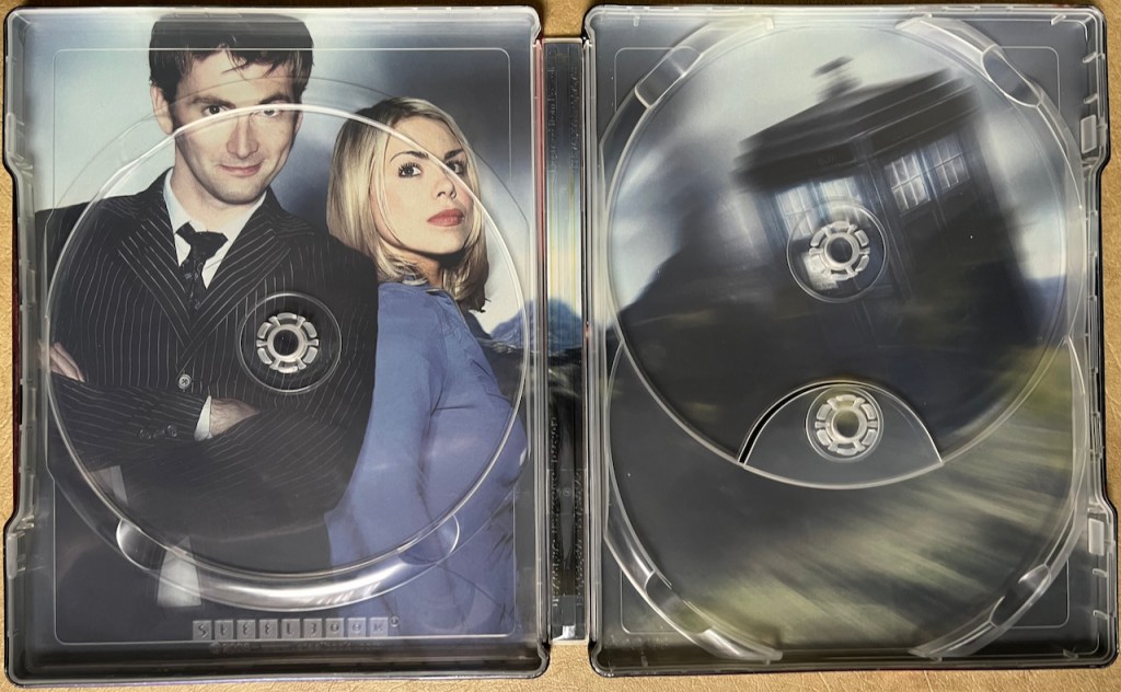 The interior spread of the Doctor Who Series 2 Blu-ray steelbook, behind where the discs are stored. The left side shows a photo of David Tennant's Doctor in a pinstripe suit and Rose in a blue top, while the right side shows a blurry image of the Tardis in flight.