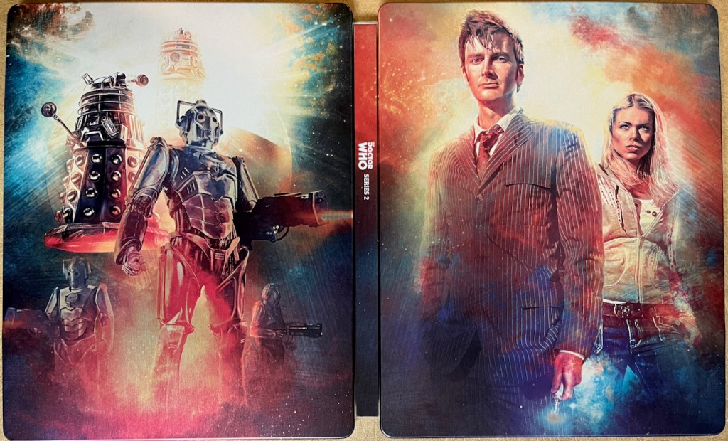 The full cover spread artwork for the Doctor Who Series 2 Blu-ray steelbook. The front cover shows David Tennant as the Doctor, wearing a pinstripe suit with bedraggled hair and his sonic screwdriver in his hand, alongside Rose who's wearing a casual jacket and jeans. Meanwhile the back cover shows Cybermen holding large guns and Daleks flying above them.