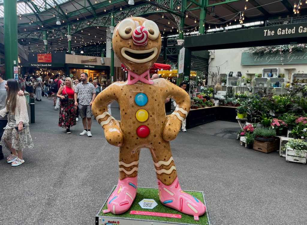Gingerbread Morph, a statue of Morph in Borough Market that looks like he's made out of gingerbread. He has a swirling red and white pattern on his nose, a pink neck tie, 3 big buttons in blue, yellow and red down his chest, pairs of wavy white lines across his wrists and lower legs, and pink shoes.
