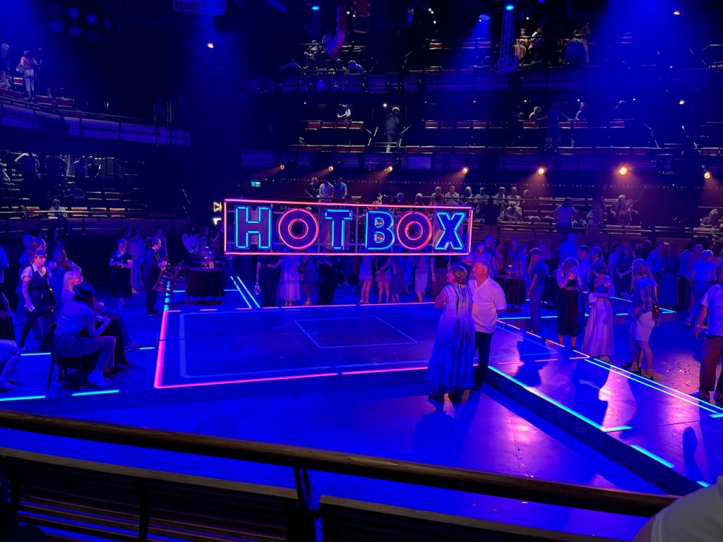 The stage for Guys and Dolls at the Bridge Theatre during the interval, completely covered in blue light. Audience members are gathered around slightly raised platforms, that form walkways leading from each corner of the stage to a central larger platform. Just above the platform is a neon sign that says Hot Box, with the letters in blue light apart from the letter O in each word in red, and a red lit border all around the name.