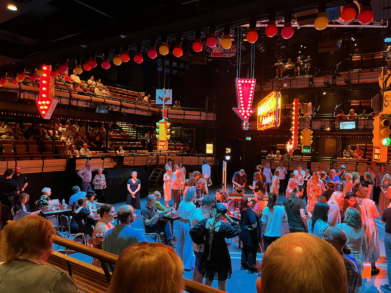 A view of the left side of the stage area, a huge square space in the middle of the auditorium, for Guys And Dolls. The stage floor is full of audience members, with a few sitting on chairs, but most are standing. Other audience members are on elevated seating around the edge of the standing area, looking down at it. Hanging from the ceiling above the stage floor are a few neon signs, including an arrow pointing downwards next to a bright sign for Mindy's restaurant. There is also a set of American traffic lights with a lit sign beneath that changes from Walk to Don't Walk.