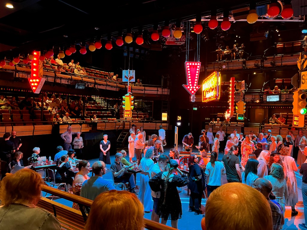 A view of the left side of the stage area, a huge square space in the middle of the auditorium, for Guys And Dolls. The stage floor is full of audience members, with a few sitting on chairs, but most are standing. Other audience members are on elevated seating around the edge of the standing area, looking down at it. Hanging from the ceiling above the stage floor are a few neon signs, including an arrow pointing downwards next to a bright sign for Mindy's restaurant. There is also a set of American traffic lights with a lit sign beneath that changes from Walk to Don't Walk.