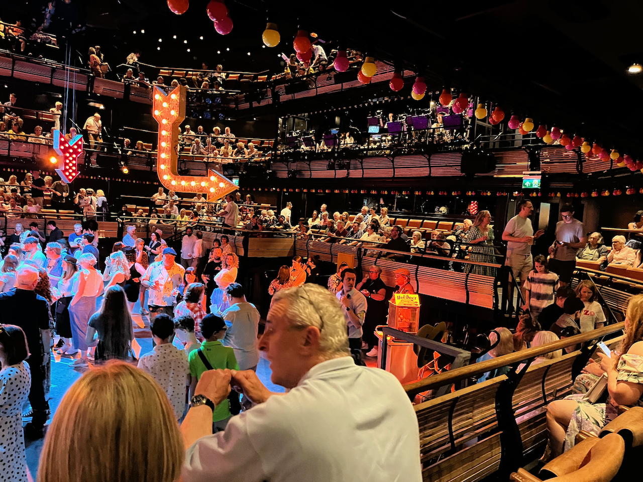A view of the right side of the stage area, a huge square space in the middle of the auditorium, for Guys And Dolls. The stage floor is full of standing audience members, while others sit in elevated rows of seats around the edge of the stage area, looking down on it. A guy selling hot dogs can also be seen on the stage floor, while a couple of brightly lit arrows hang down from the ceiling. Above the audience members seated on the right edge of the stage space, a long rectangular opening in the wall shows the 14-piece band who play live during the show.