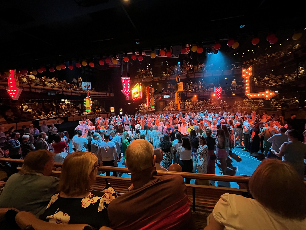 The stage for Guys and Dolls at the Bridge Theatre, a huge square space with seating on all 4 sides. In the space, hundreds of audience members are gathered beneath New York style traffic lights and neon signs that hang from the ceiling.