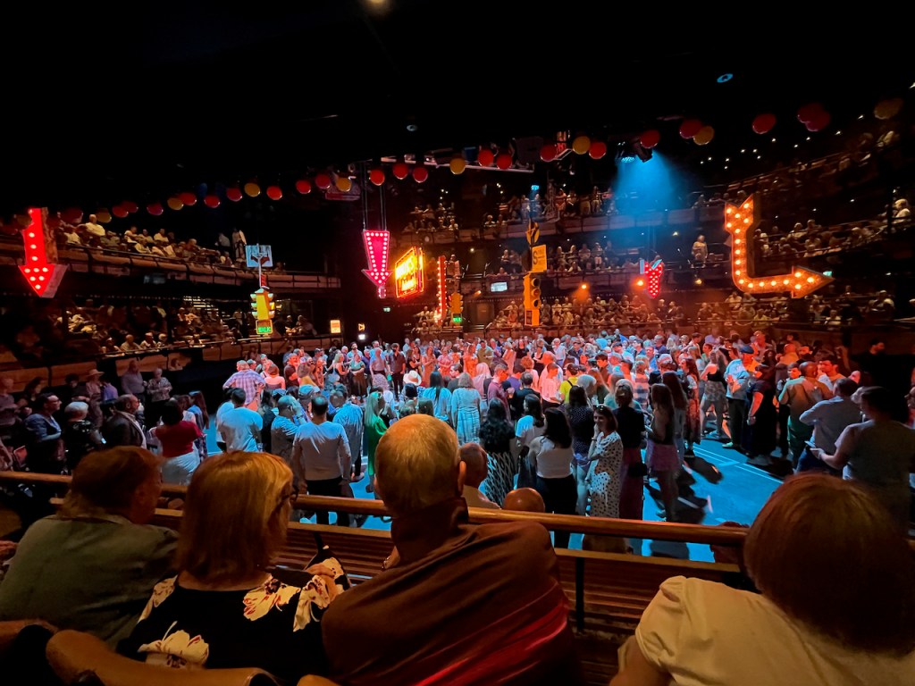 The stage for Guys and Dolls at the Bridge Theatre, a huge square space with seating on all 4 sides. In the space, hundreds of audience members are gathered beneath New York style traffic lights and neon signs that hang from the ceiling.