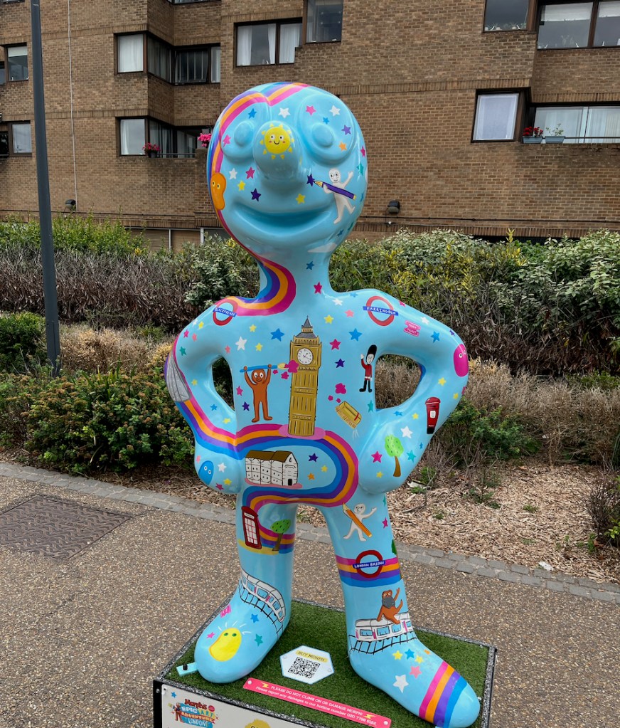I Spy Morph, a light blue statue of Morph covered in a variety of images, including rainbows, colourful stars, Big Ben, Tube station roundels, trains, Morph holding paintbrushes and pencils, Morph dressed as a royal guard, trees, a red phone box, a red post box, and more.