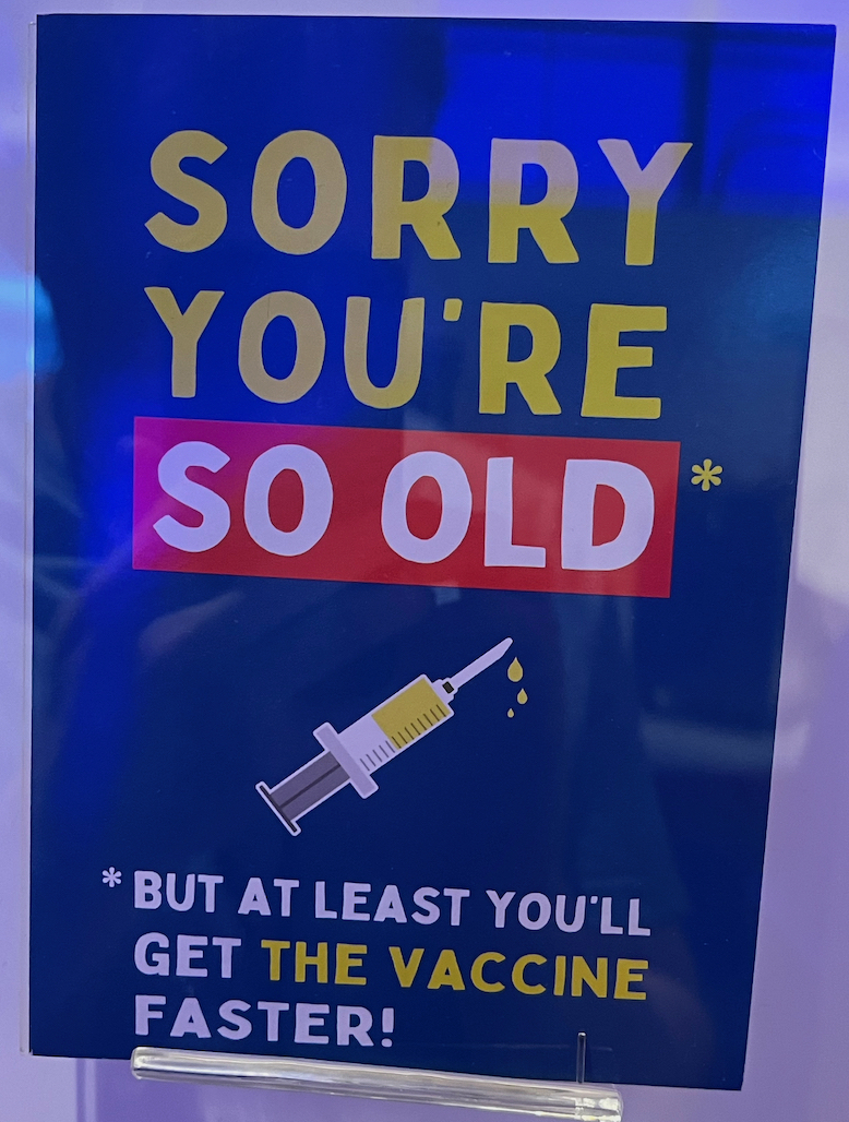 A greeting card that says Sorry you're so old, but at least you'll get the vaccine faster! The text is split in the middle by a drawing of a small syringe spurting out liquid.