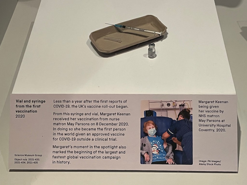 A syringe and a small glass vial, used when Margaret Keenan became the first person to receive an approved vaccine outside a clinical trial. The panel below shows a photo of her receiving the vaccination while wearing a blue top with a Christmassy penguin on it.