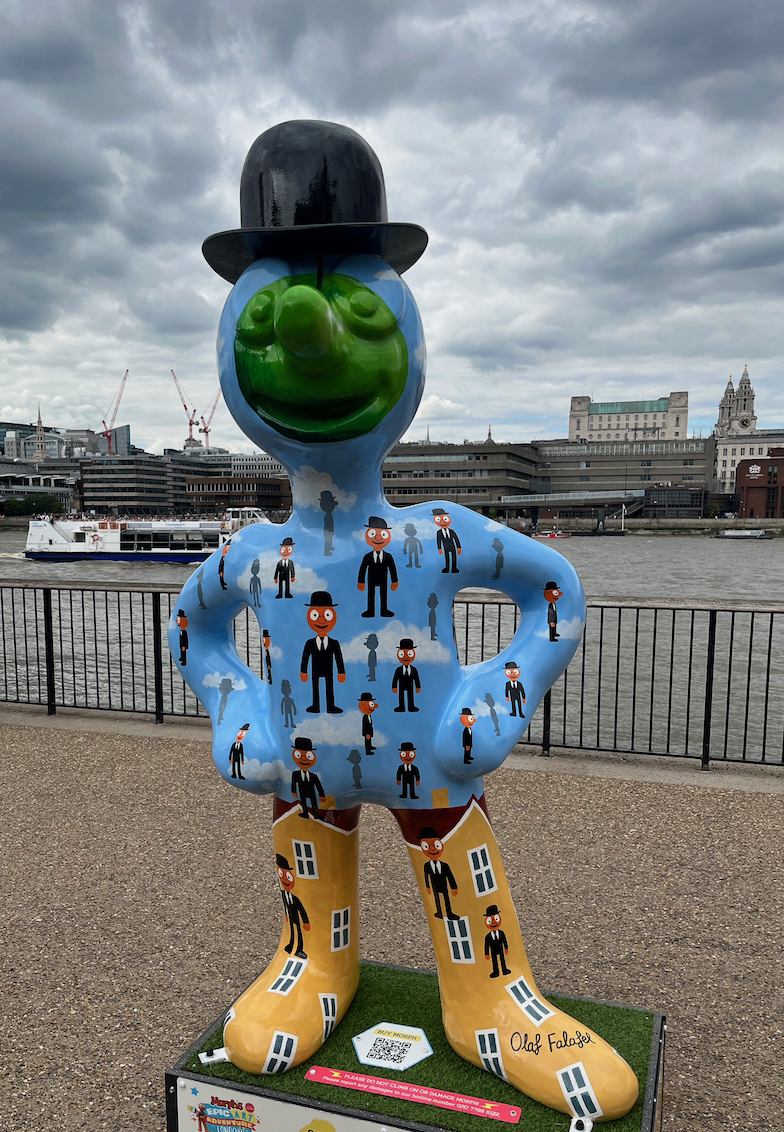 It’s Raining Morphs, Hallelujah, a statue of Morph whose light blue body is covered in images of lots of smart Morphs wearing black suits and black bowler hats, who look like they're floating in the sky as there are clouds in the background. The Morph statue is also wearing a black bowler hat, has a dark green circle covering his facial features, and his boots have the design of houses with windows, with more smart Morphs floating in front of them.