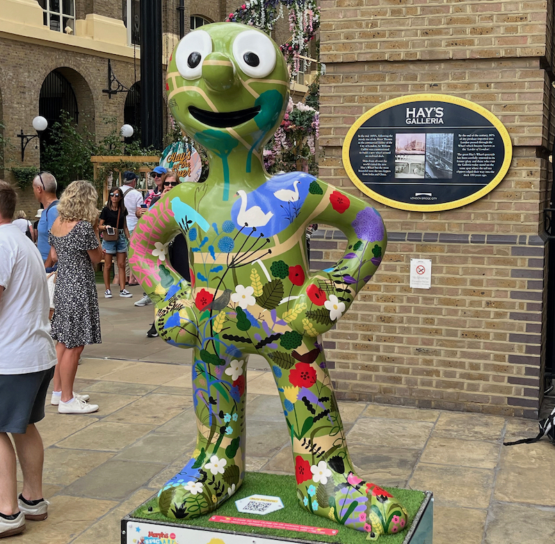 London Parklife, a statue of Morph covered in a painted scene of a green park with swans in a lake and various flowers.