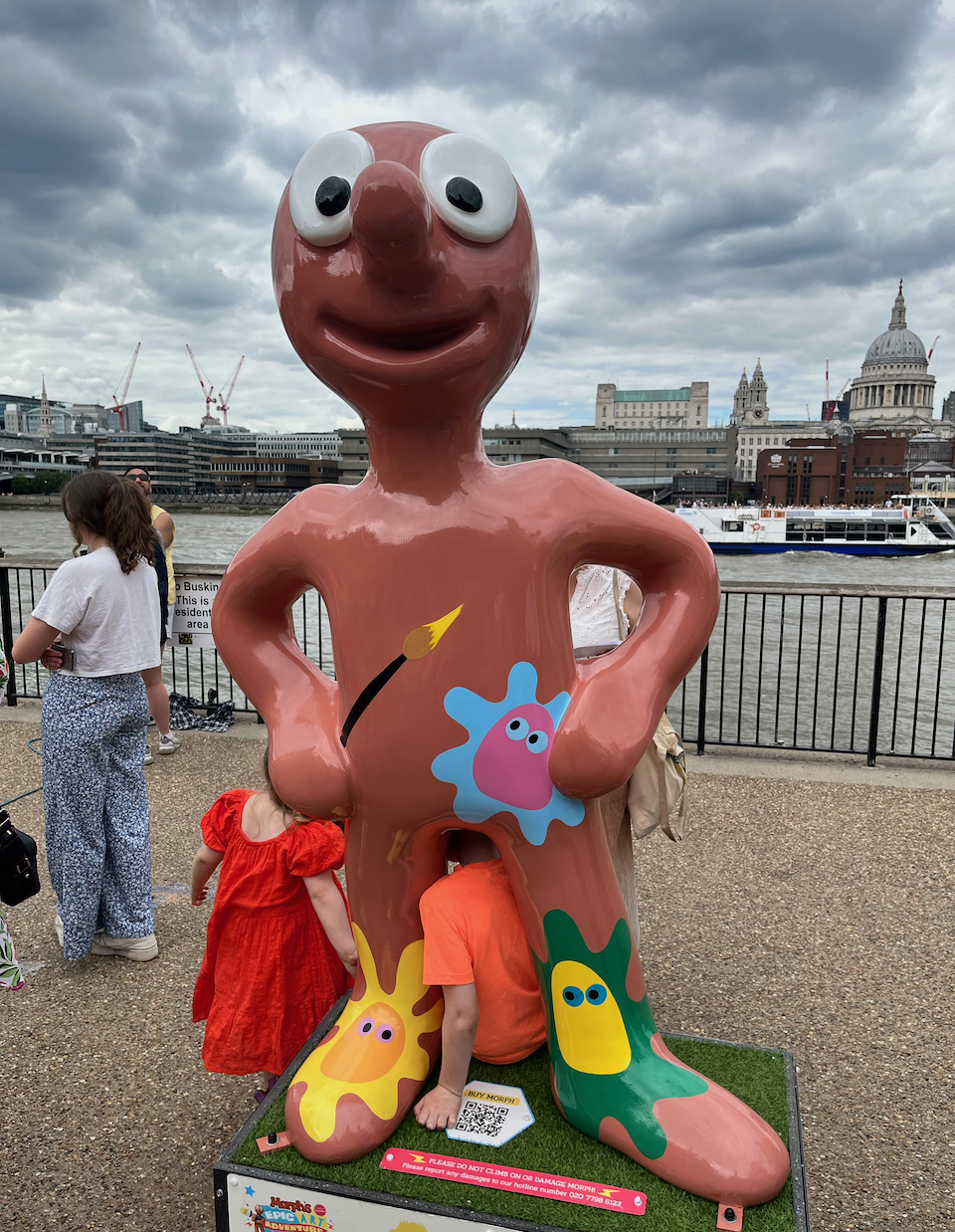Mega Messy Morph, a statue of a red clay Morph holding a paintbrush, with a few splodges of paint on his body. On his lower chest is a pink blob with eyes, surrounded by a messy blue border. His right foot has an orange blob with eyes, and a messy yellow border. And his left leg has a yellow blob with blue eyes and no border.