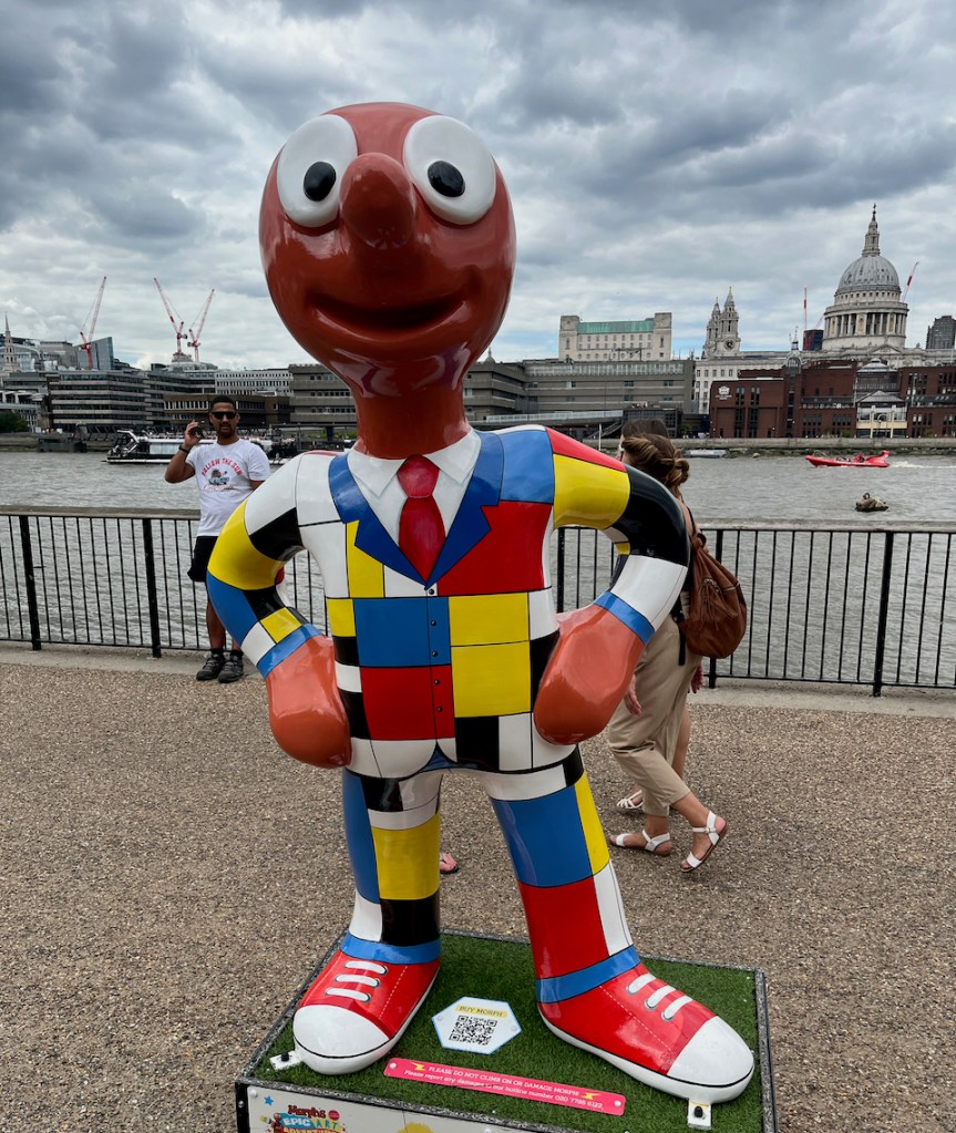 Mondrian Morph, a statue of Morph wearing a suit covered in a random combination of squares and rectangles in white, black, blue, red and yellow colours. Beneath the jacket is a red tie and a white shirt. On his feet he has red shoes with white laces and white toecaps. St Paul's Cathedral can be seen to the right in the background.